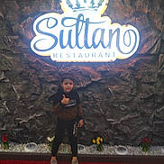 Amir participe au concours pour gagner de l'argent avec cette photo : child, boy, thumbs_up, restaurant_sign, stone_wall, flowers, indoor, casual, dark_clothing, standing, smile, decor, glowing_sign, red_flowers, yellow_flowers, white_flowers, floor, pose, portrait, lighting