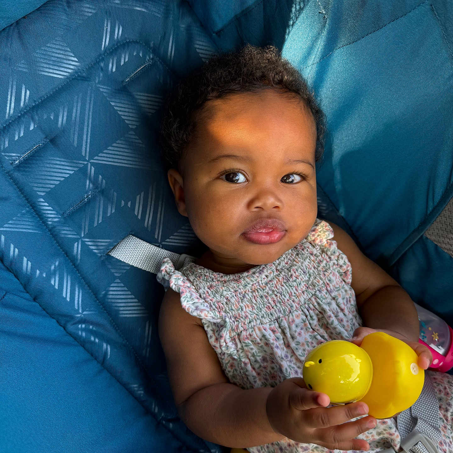 Noor Nala is registered to the contest to win money with this photo: baby, blue_cushion, child, clothing, curly_hair, cute, face, floral_dress, hand, happiness, indoors, infant, playful, portrait, seat, sitting, smiling, soft_light, toy, yellow_duck