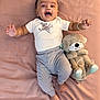 Yunay is registered to the contest to win money with this photo: baby, child, smiling, happy, plush_toy, teddy_bear, onesie, striped_pants, bracelet, bed, blanket, soft_toy, infant, cute, portrait, lying_down, indoors, person, face, hand