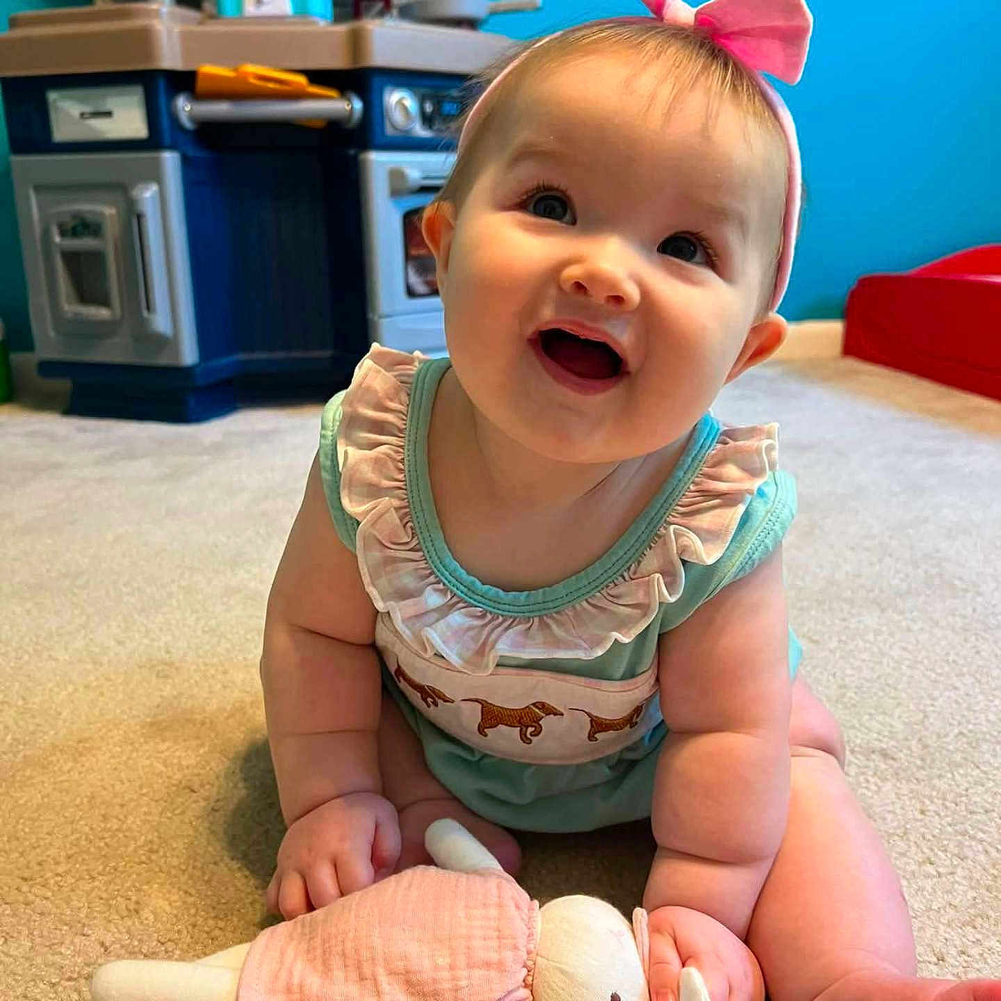 Eleonor is registered to the contest to win money with this photo: baby, child, infant, pink_bow, toy, stuffed_animal, carpet, play_kitchen, happy, smiling, indoors, cute, person, toddler, headband, blue_wall, floor, clothing, play, childhood