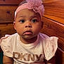 baby, child, chubby_arms, closeup, clothing, curious_expression, cute, dkny_shirt, face, flower, headwear, indoors, infant, person, pink_headband, portrait, sitting, skin, wooden_background, young_child