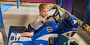 Elio participe au concours pour gagner de l'argent avec cette photo : blue, boy, child, floor, fun, hand_on_handlebar, indoor, kid, lighting, motorcycle, netting, padded_walls, play, play_area, police_ride, shorts, smiling, toy_vehicle, tshirt, young
