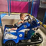 blue, boy, child, floor, fun, hand_on_handlebar, indoor, kid, lighting, motorcycle, netting, padded_walls, play, play_area, police_ride, shorts, smiling, toy_vehicle, tshirt, young