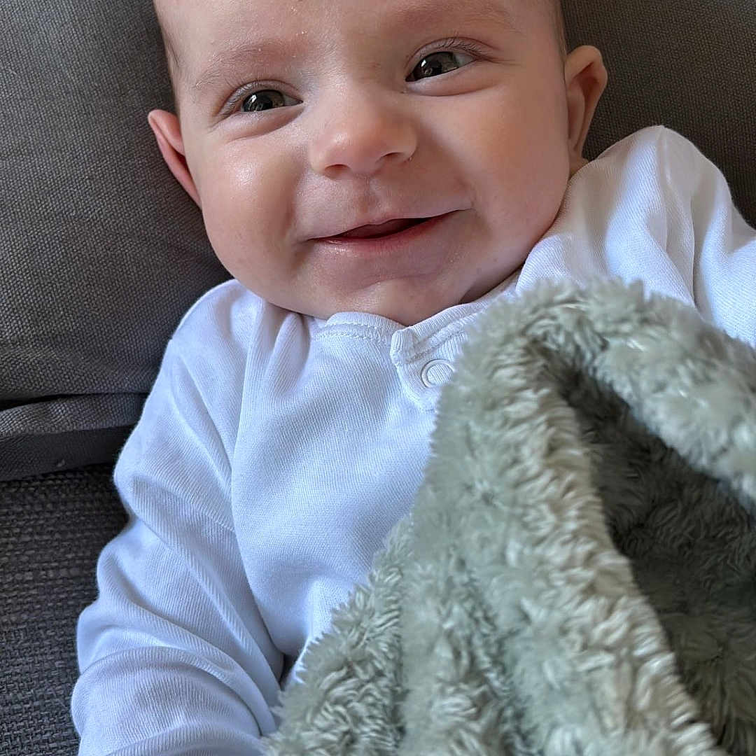 Theo is registered to the contest to win money with this photo: baby, blanket, clothing, couch, cushion, face, furniture, happy, head, homedecor, laughing, linen, newborn, person, photography, portrait, quilt, sitting, smile, towel