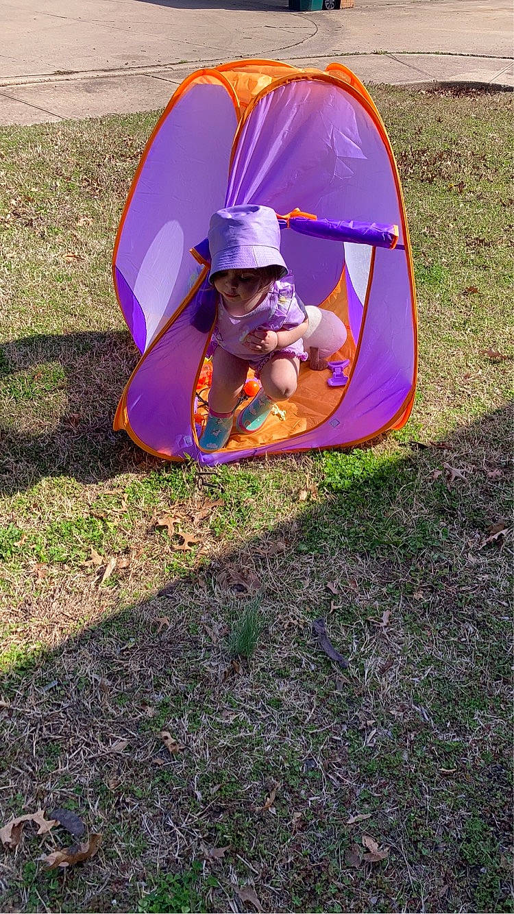 Isabella is registered to the contest to win money with this photo: city, comfort, fun, grass, grassland, hat, landscape, leisure, magenta, outdoor_play_equipment, outdoor_recreation, person, plant, purple, recreation, shade, sitting, tent, tints_and_shades, toddler