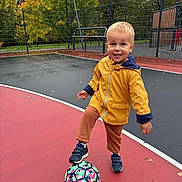 Tom participe au concours pour gagner de l'argent avec cette photo : toddler, child, soccer_ball, yellow_jacket, outdoor, playground, autumn, trees, fence, court, sport, smiling, shoes, pants, recreation, activity, fun, daylight, park, happy