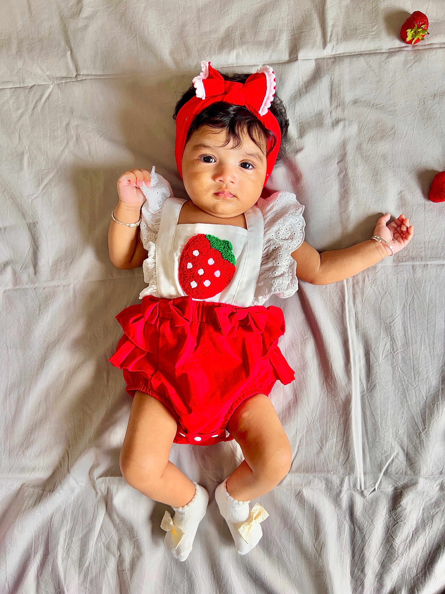 Raikishori is registered to the contest to win money with this photo: baby, infant, red_headband, strawberry_outfit, white_socks, bow, lying_down, cute, soft_sheet, fruit, strawberry, adorable, child, person, portrait, indoors, colorful, fashion, baby_clothes, headwear