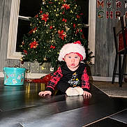 Amira is registered to the contest to win money with this photo: baby, christmas_tree, santa_hat, holiday_decorations, festive_clothing, crawling, floor, indoors, christmas_lights, bucket, ghost_illustrations, window, wall, chair, carpet, red_bows, ornaments, plaid, person, cute