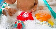 Felix is registered to the contest to win money with this photo: baby, child, bath, bathtub, bubbles, foam, water, bath_time, toy, colorful_toys, smiling, happy, wet, infant, portrait, indoor, warm_lighting, bokeh, blue_tub, soap