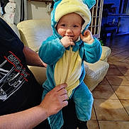 Tyler participe au concours pour gagner de l'argent avec cette photo : adult, baby, blue, child, costume, cute, floor, furniture, hand, hood, indoor, living_room, onesie, person, playful, plush, smile, sofa, tile_floor, yellow