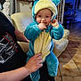 adult, baby, blue, child, costume, cute, floor, furniture, hand, hood, indoor, living_room, onesie, person, playful, plush, smile, sofa, tile_floor, yellow