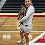 person, dog, french_bulldog, basketball_court, sports_uniform, white_shoes, smiling, indoor, bleachers, wood_floor, athlete, pet, holding, young_woman, shorts, socks, team, gym, portrait, happy