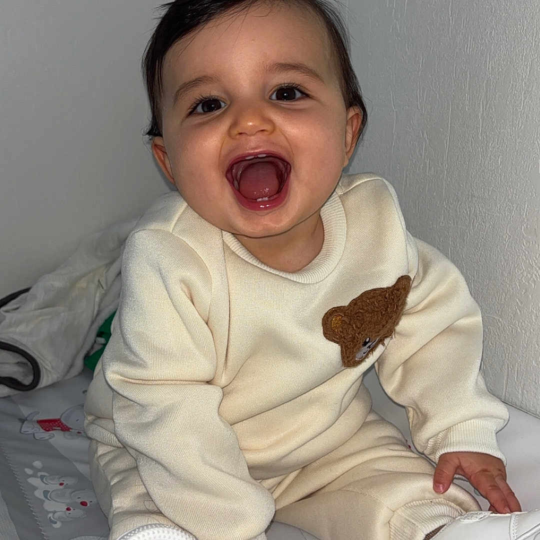 Tilyano a rejoint le concours — aidez-le/la à gagner de superbes lots ! baby, cheerful, child, clothing, cream_outfit, cute, face, footwear, happy, head, indoors, infant, patterned_mat, person, portrait, sitting, smiling, teddy_bear_patch, white_shoes, young_child