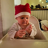 Ellie a rejoint le concours — aidez-le/la à gagner de superbes lots ! baby, blanket, child, christmas, cozy, decorations, face, fairy_lights, hands, high_chair, holiday, indoor, infant, pinecones, red_hat, santa_hat, table, warm_light, white_clothing, wooden_shelf
