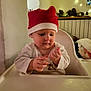 baby, blanket, child, christmas, cozy, decorations, face, fairy_lights, hands, high_chair, holiday, indoor, infant, pinecones, red_hat, santa_hat, table, warm_light, white_clothing, wooden_shelf