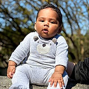 Idriss participe au concours pour gagner de l'argent avec cette photo : baby, infant, child, person, face, portrait, sitting, outdoors, trees, bokeh, sunlight, onesie, plush_bear_design, hands, feet, stone_surface, cheeks, hair, curious_expression, soft_texture