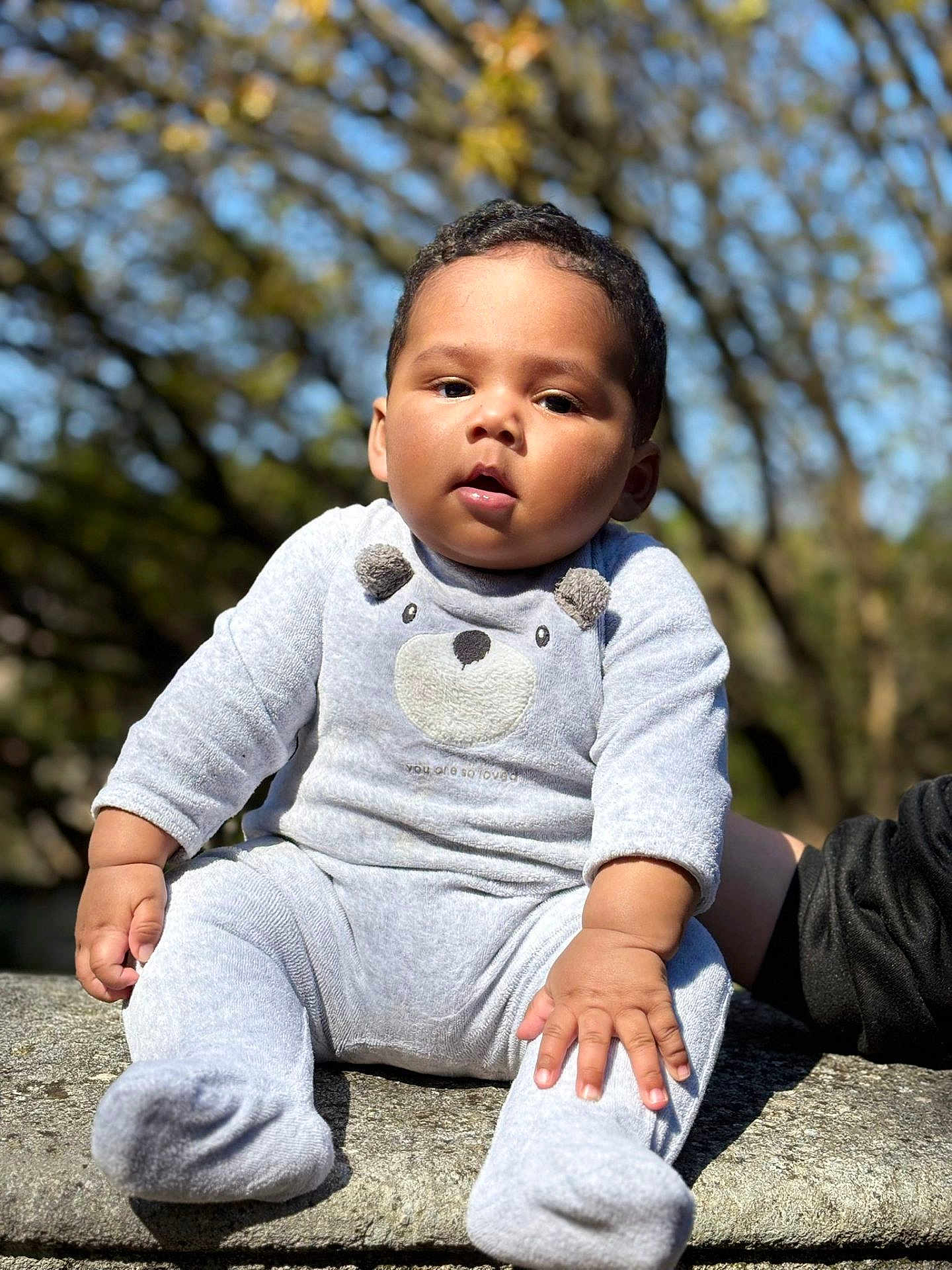 Idriss participe au concours pour gagner de l'argent avec cette photo : baby, infant, child, person, face, portrait, sitting, outdoors, trees, bokeh, sunlight, onesie, plush_bear_design, hands, feet, stone_surface, cheeks, hair, curious_expression, soft_texture