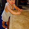 toddler, child, baby, standing, barefoot, white_outfit, portrait, indoors, table, furniture, television, shelves, home, brown_hair, facial_expression, looking_at_camera, arms, hands, legs, photograph