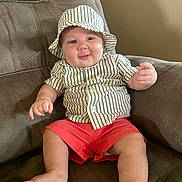 Grayson is registered to the contest to win money with this photo: baby, child, infant, hat, striped_clothing, shorts, red_shorts, shirt, striped_shirt, armchair, furniture, smile, happy, cute, indoors, person, plush, relaxed, comfortable, portrait