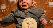 Leonidas is registered to the contest to win money with this photo: baby, blanket, child, clothing, cozy, cute, face, hand, happy, indoor, infant, knitwear, lying_down, pattern, portrait, smiling, sun, two_months, warm_colors, wooden_sign
