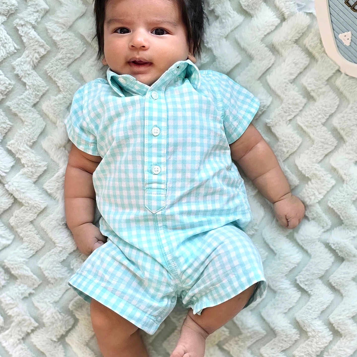 Advik is registered to the contest to win money with this photo: baby, blanket, blue_clothing, checkered_pattern, child, chubby_arms, chubby_legs, cute, dark_hair, face, human, indoors, infant, lying_down, one_person, portrait, romper, smiling, soft_texture, textured_blanket
