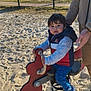 child, toddler, playground, spring_rocker, sand, fence, lake, trees, adult_hand, blue_sky, jacket, pants, shoes, outdoor, daylight, play, person, park, toy, nature