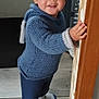 toddler, child, smile, blue_sweater, socks, standing, door_frame, wooden_door, floor, indoor, person, face, happy, hair, hand, cat_food_bag, background, clothing, home, young_child