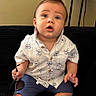 baby, child, sitting, indoors, black_couch, white_shirt, blue_shorts, sneakers, sunglasses, short_hair, mohawk, expression, face, person, home_decor, shelf, casual_clothing, cute, fashion, portrait