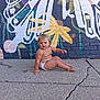 toddler, child, diaper, sidewalk, concrete, brick_wall, mural, colorful_paint, outdoor, barefoot, expression, sitting, playful, young_child, urban, street_art, casual, skin, person, daylight