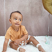 Blair-Ryser is registered to the contest to win money with this photo: background, balloon, boy, casual_clothing, child, curious_expression, cute, fluffy_rug, gold_balloon, indoor, orange_shirt, playful, portrait, sitting, smiling, socks, soft_lighting, striped_shorts, toddler, white_rug