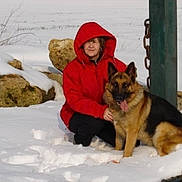 Sacco participe au concours pour gagner de l'argent avec cette photo : child, german_shepherd, dog, snow, winter, outdoor, red_coat, hood, sitting, portrait, pet, animal, snowy_landscape, rocks, wooden_post, chain, cold_weather, smiling, family_pet, playful