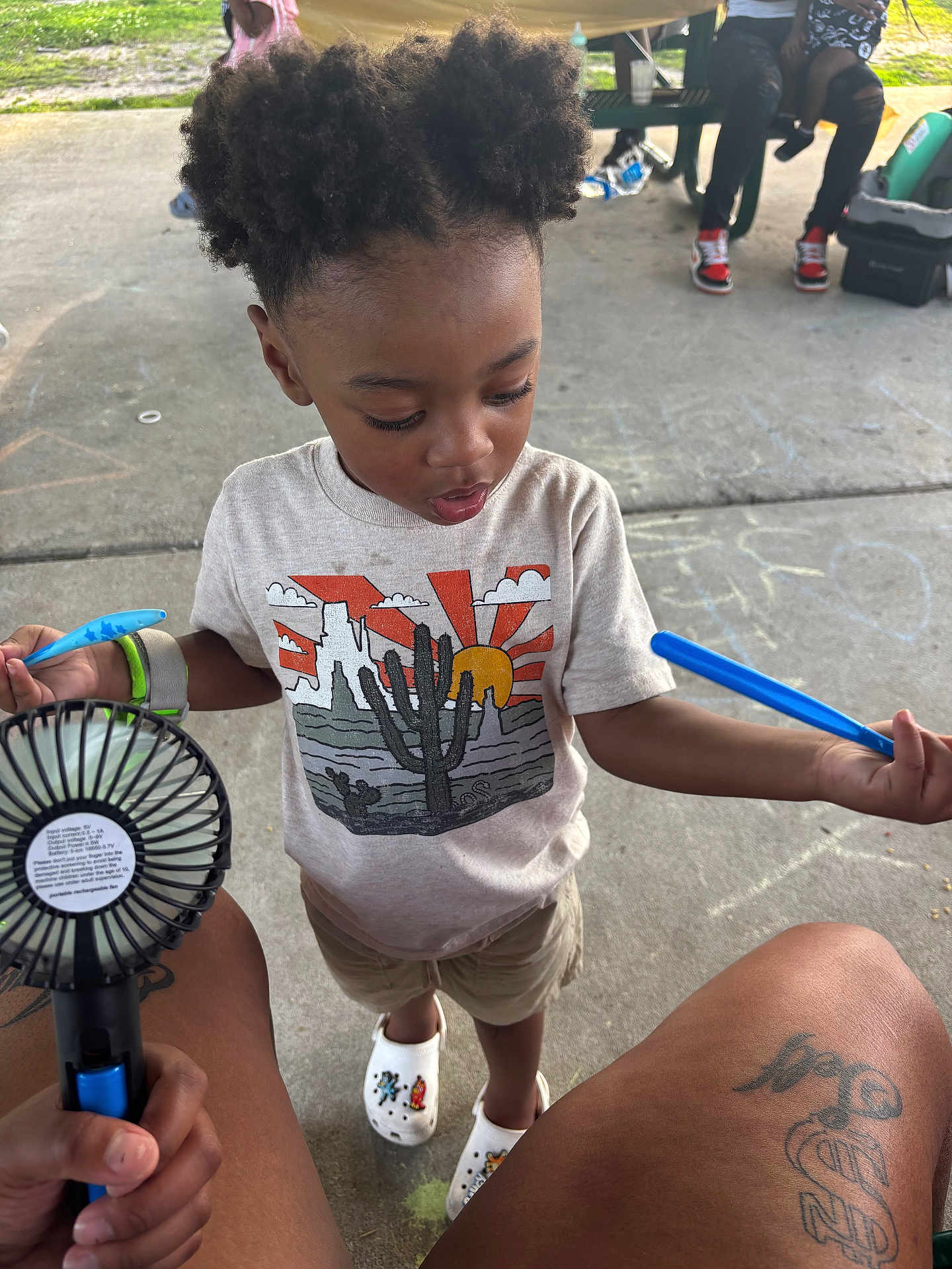 Kashmir is registered to the contest to win money with this photo: child, toddler, hair, tshirt, cactus, crocs, chalk, concrete, fan, legs, person, handheld_fan, outdoor, picnic_table, drawing, shoes, shorts, tattoo, relaxing, summer