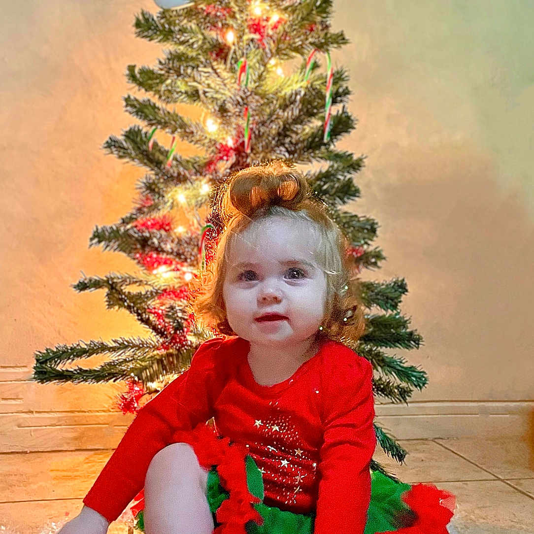 Liliana joined the competition — help win amazing prizes! accessories, child, christmas, christmasdecorations, clothing, conifer, face, female, festival, girl, head, lighting, pants, person, photography, plant, portrait, pottedplant, sitting, tree