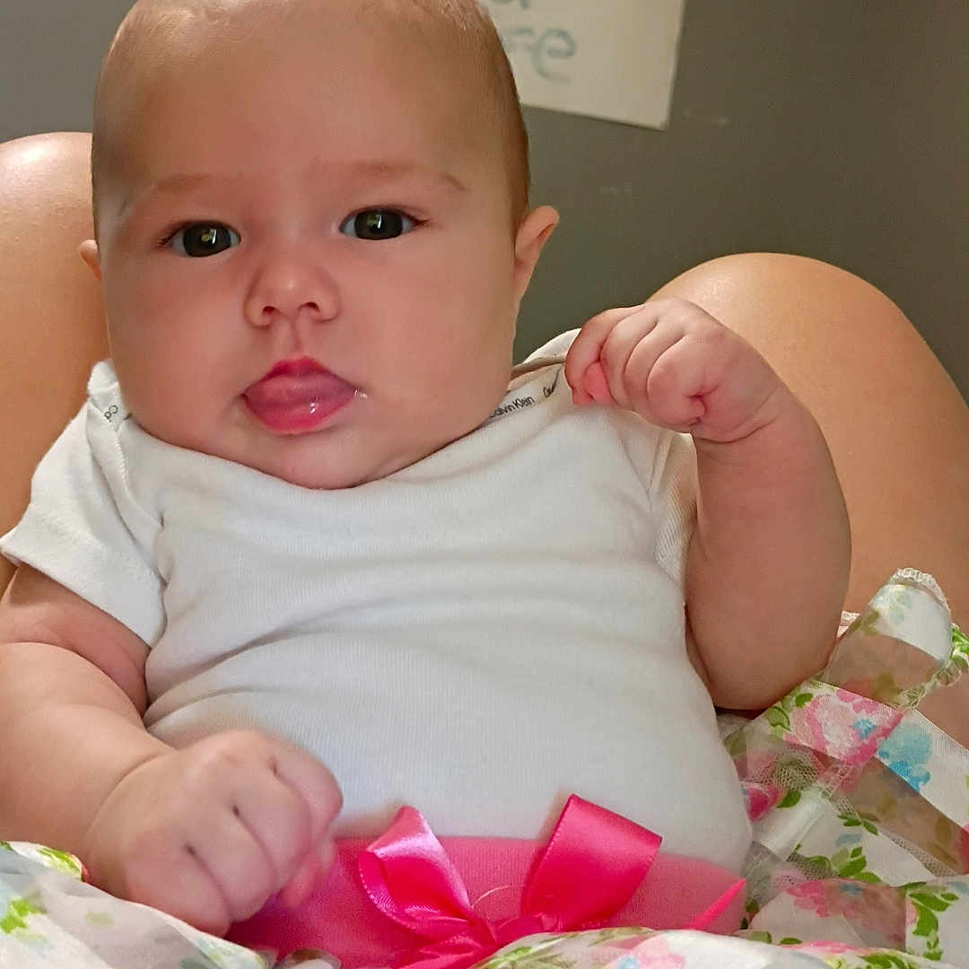 Mia joined the competition — help win amazing prizes! baby, child, infant, white_onesie, floral_skirt, pink_bow, tongue_out, cute, chubby_cheeks, indoor, person, portrait, sitting, handmade_sign, colorful, young_child, skin, face, curious_expression, background