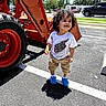 asphalt, blue_shoes, boy, cars, child, curly_hair, machine, outdoor, parking_lot, person, road_marking, shadow, smiling, sunny_day, tan_shorts, toddler, tractor, trees, vehicle, white_tshirt