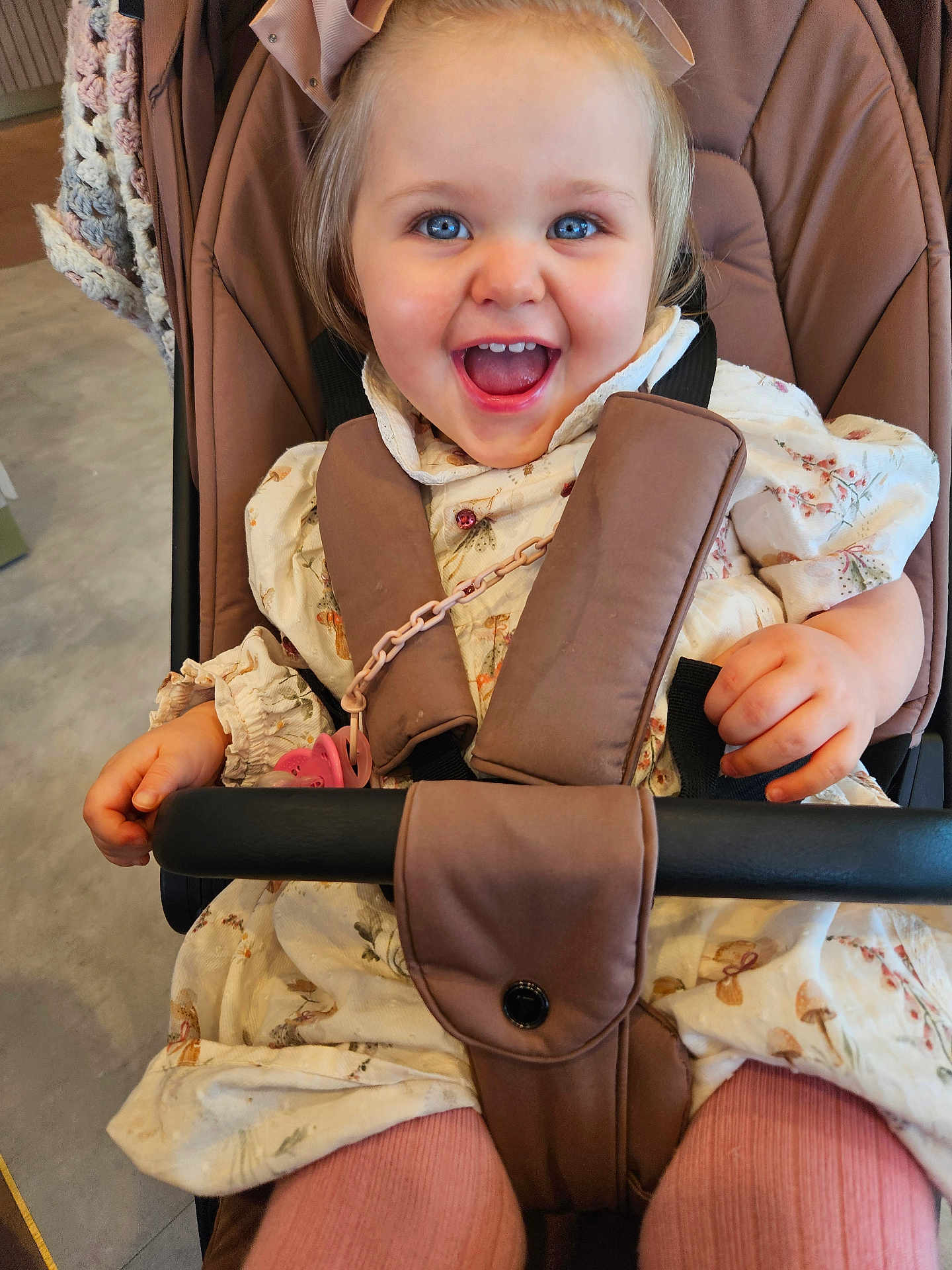 Nyla is registered to the contest to win money with this photo: child, toddler, smiling, happy, stroller, baby, blue_eyes, dress, pink_tights, seatbelt, indoor, cute, hand, face, person, infant, clothing, baby_accessory, joy, young