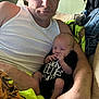 man, baby, infant, person, couch, clothing, white_tank_top, black_onesie, indoor, casual, holding, relaxing, home, blanket, denim_jacket, arm, face, skin, furniture, portrait