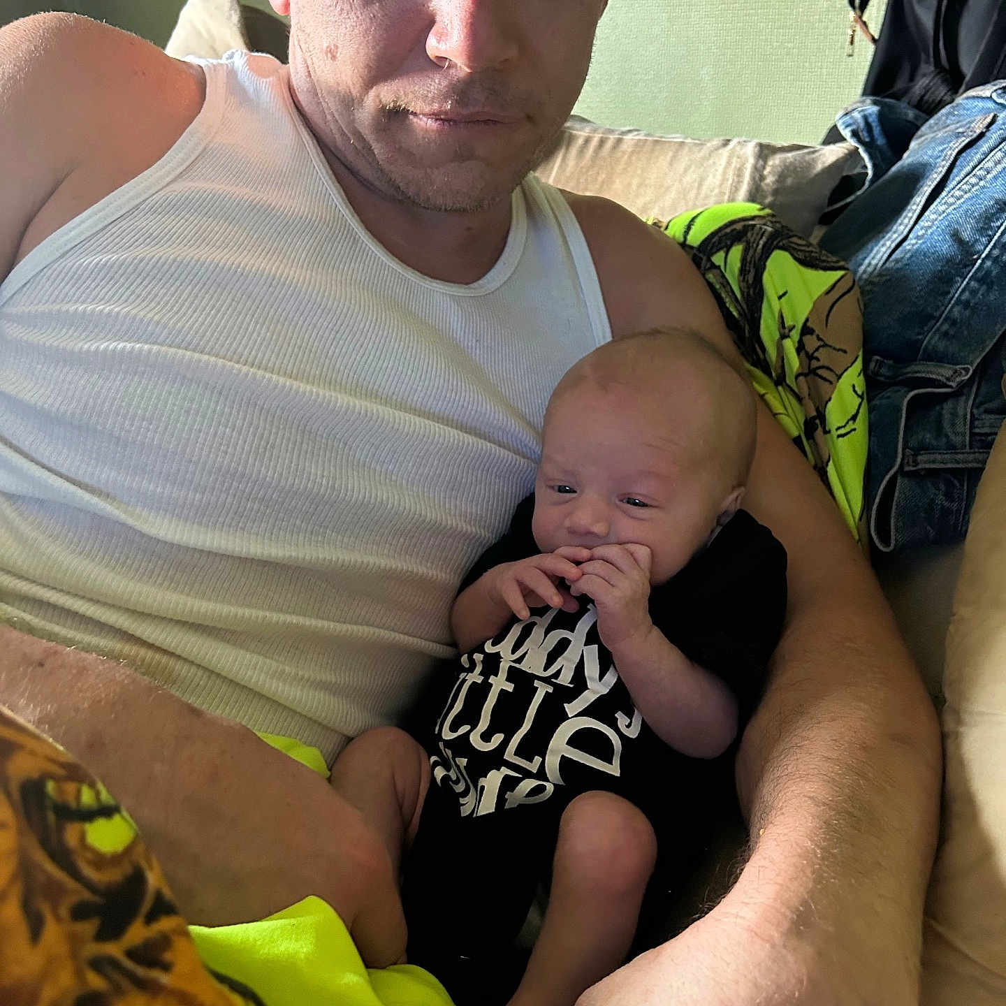 Lusten Holloway is registered to the contest to win money with this photo: arm, baby, black_onesie, blanket, casual, clothing, couch, denim_jacket, face, furniture, holding, home, indoor, infant, man, person, portrait, relaxing, skin, white_tank_top