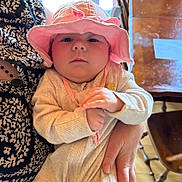 Maeve joined the competition — help win amazing prizes! baby, infant, pink_hat, holding, adult_hand, patterned_clothing, indoor, wooden_table, wooden_chair, serious_expression, close_up, cozy_outfit, curious, child, person, portrait, seated, face, hat, soft_lighting