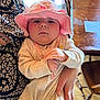 baby, infant, pink_hat, holding, adult_hand, patterned_clothing, indoor, wooden_table, wooden_chair, serious_expression, close_up, cozy_outfit, curious, child, person, portrait, seated, face, hat, soft_lighting
