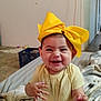 baby, child, yellow_bow, heart_necklace, smiling, happy, indoor, blanket, jewelry, cute, portrait, sitting, infant, person, clothing, accessory, earrings, room, floor, expression