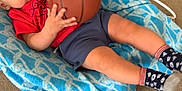 Giovanni is registered to the contest to win money with this photo: baby, basketball, blue_blanket, carpet, child, clothing, curious, floor, indoor, infant, legs, person, red_shirt, socks, small_basketball, toy, young_child, relaxing, patterned_blanket, hand