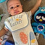 baby, child, carpet, plush_toy, blue_bunny, toy_truck, onesie, bib, easter, egg_pattern, indoor, floor, cute, infant, smiling, lying_down, playtime, colorful, young_child, happy