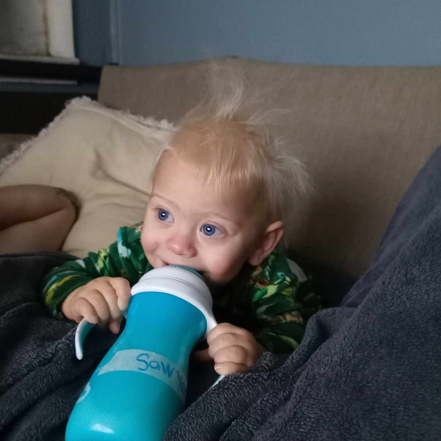 Sawyer is registered to the contest to win money with this photo: baby, child, infant, smiling, blue_eyes, blond_hair, sippy_cup, drinking, hands, green_clothing, blanket, couch, pillow, indoor, home, portrait, close_up, sitting, happy, cozy