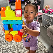 Journey-Joy is registered to the contest to win money with this photo: toddler, child, toy, blocks, train, hair_buns, indoor, carpet, television, furniture, pink_chair, doll, playroom, smiling, barefoot, purple_shirt, shorts, earrings, colorful, home