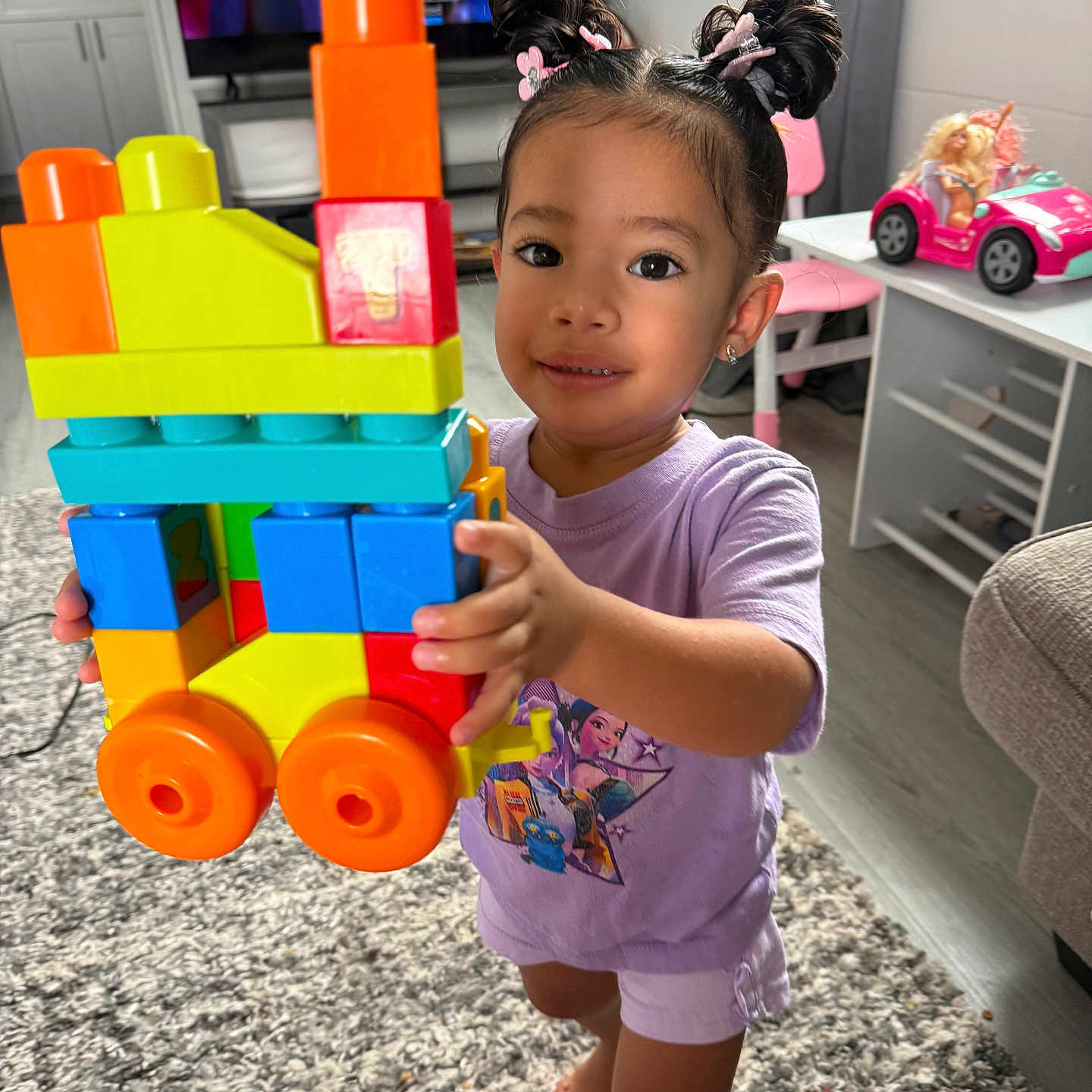 Journey-Joy is registered to the contest to win money with this photo: toddler, child, toy, blocks, train, hair_buns, indoor, carpet, television, furniture, pink_chair, doll, playroom, smiling, barefoot, purple_shirt, shorts, earrings, colorful, home