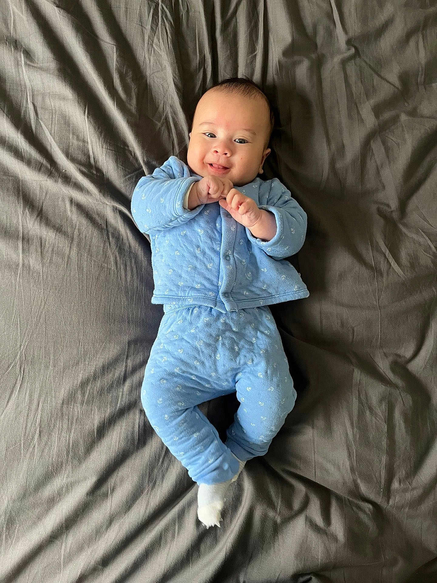 Gabriel participe au concours pour gagner de l'argent avec cette photo : baby, infant, smiling, blue_pajamas, lying_down, gray_bed_sheet, cute, portrait, indoor, one_person, hands_together, socks, cheeks, newborn, soft_texture, happy, small_hands, head, face, cozy