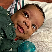 Legend is registered to the contest to win money with this photo: baby, smile, face, pacifier, green_clothing, pillow, blanket, infant, lying_down, closeup, happy, child, cute, indoors, skin, head, expression, newborn, person, portrait