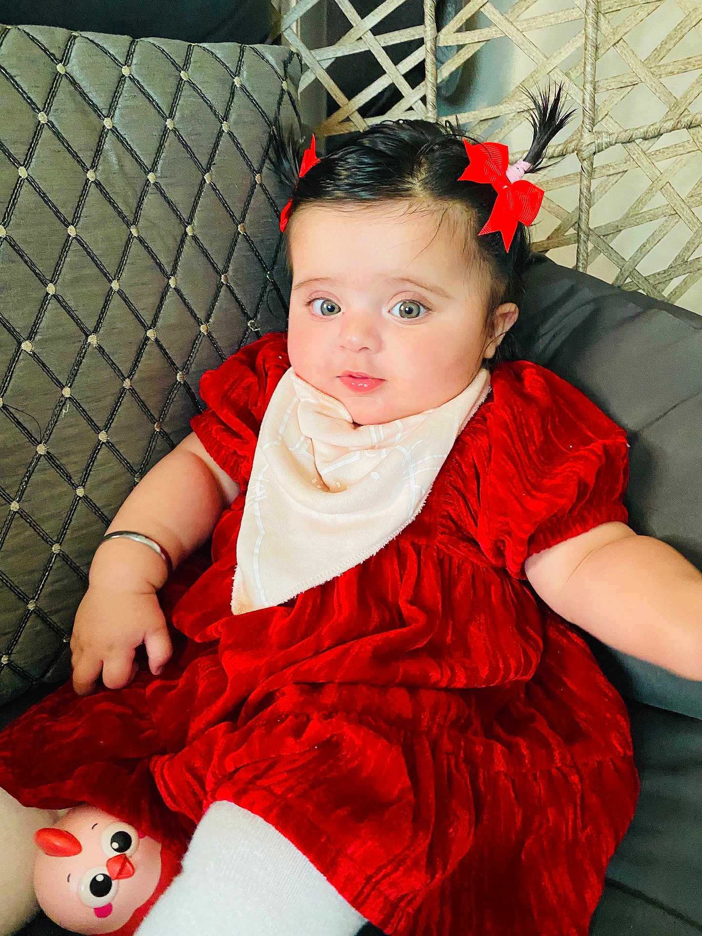Sidak is registered to the contest to win money with this photo: baby, child, red_dress, pigtails, red_bows, bib, couch, toy, pink_toy, face, smiling, indoor, cute, chubby_cheeks, person, sitting, happy, bright_eyes, furniture, plush
