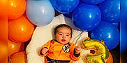 Hans is registered to the contest to win money with this photo: baby, orange_outfit, blue_balloons, orange_balloons, gold_balloon, number_3, dragon_ball_z, figurines, fluffy_blanket, birthday, celebration, child, smiling, lying_down, indoor, cute, colorful, toy_figures, decorations, happy